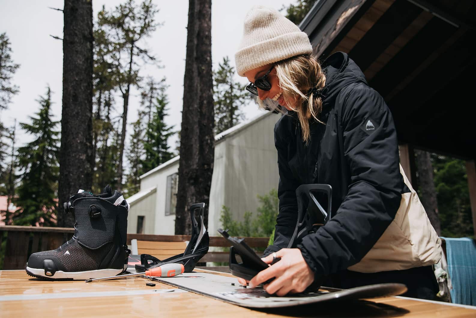 How to set up a snowboard: A woman smiles enthusiastically while adjusting snowboard bindings on an outdoor workbench.