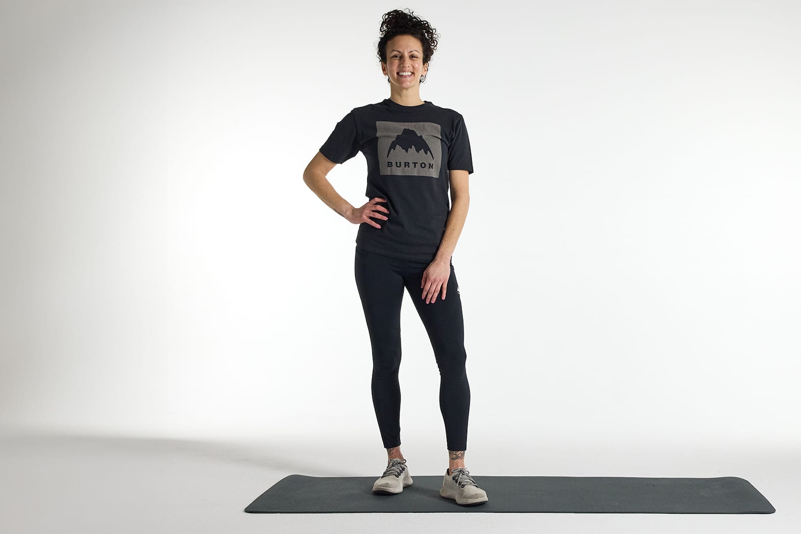 The snowboarder's workout: A woman in a Burton t-shirt and leggings standing on a yoga mat, smiling.