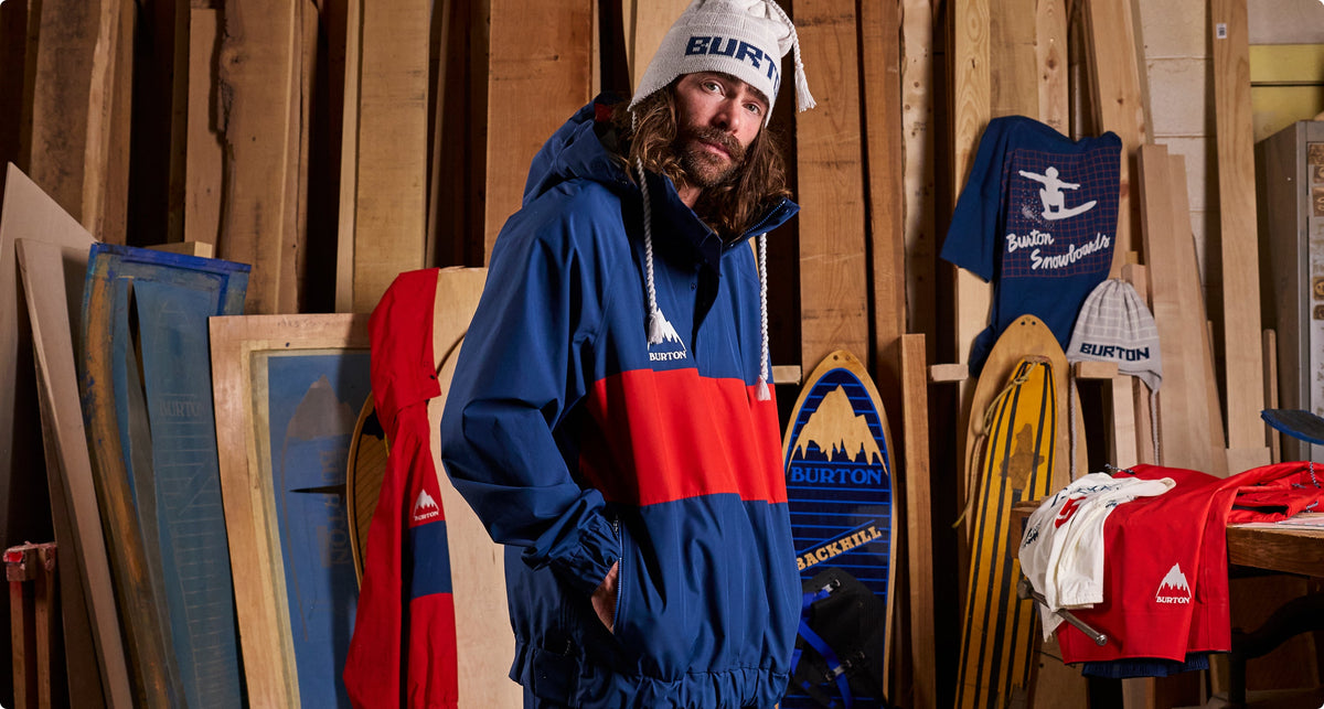 Danny Daviswearing a blue, red, and white Burton windbreaker jacket and a white beanie stands in a workshop filled with wood panels, snowboards, and other Burton apparel.