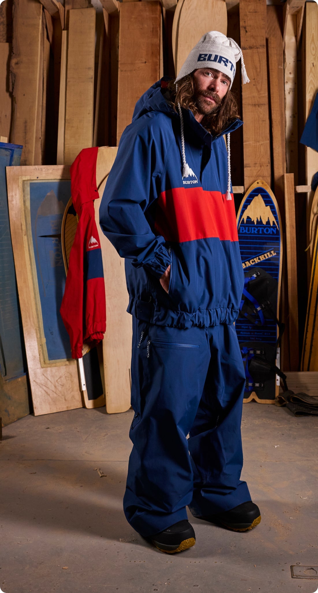 Danny Davis wearing a blue and red Burton snow jacket and matching snow pants, standing in a workshop-like environment with wood panels and snowboards in the background.