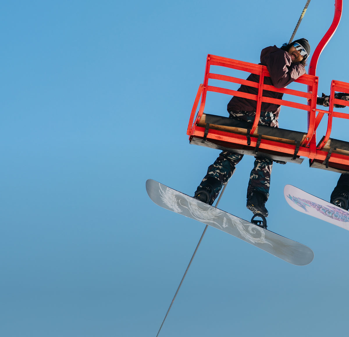 A snowboarder in a red ski lift basket against a clear blue sky, with the snowboard and part of another board visible.
