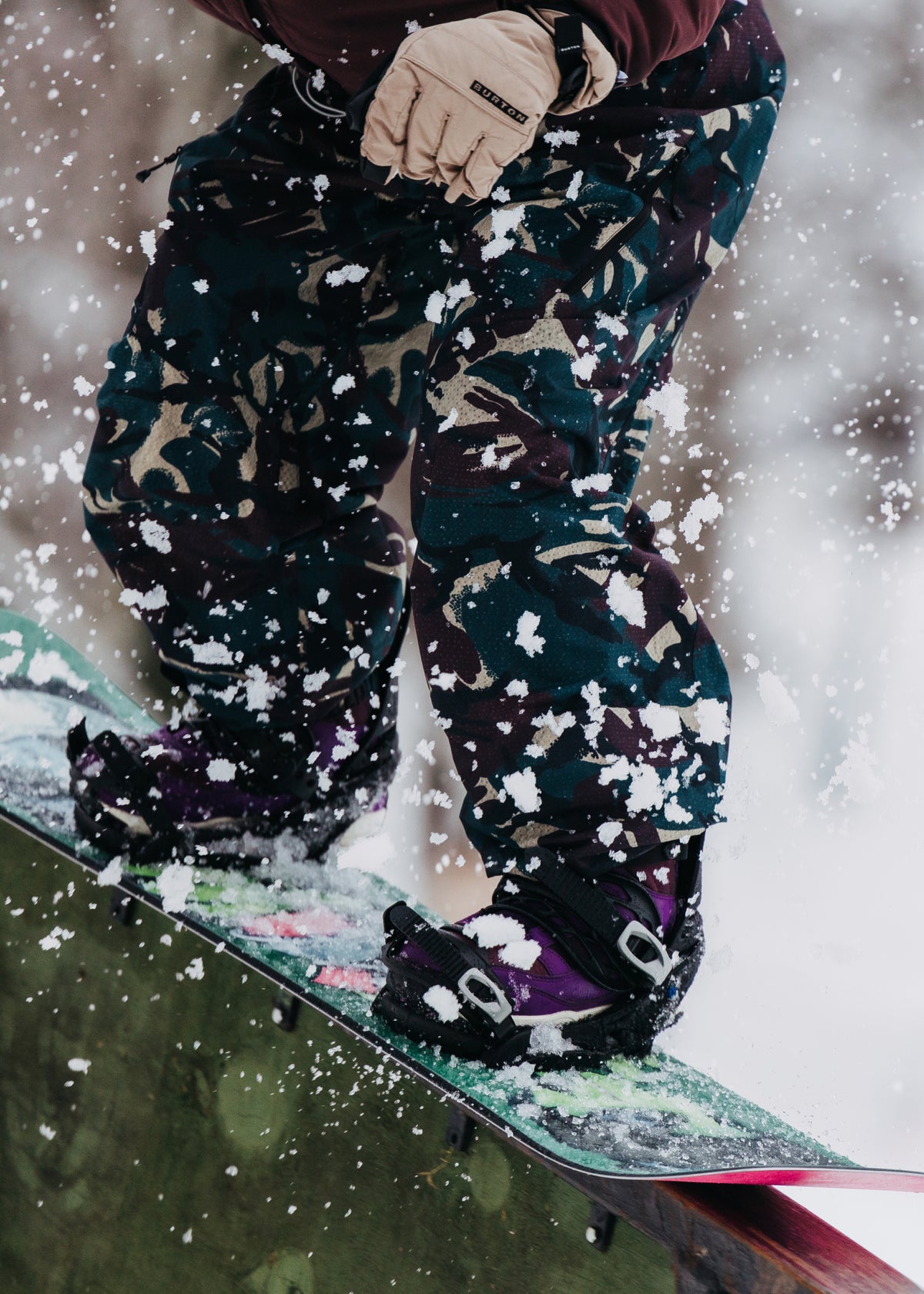 Close-up of a snowboarder's lower body wearing dark camouflage snow pants with purple bindings and a snowboard covered in snow, with snow spraying in the air.