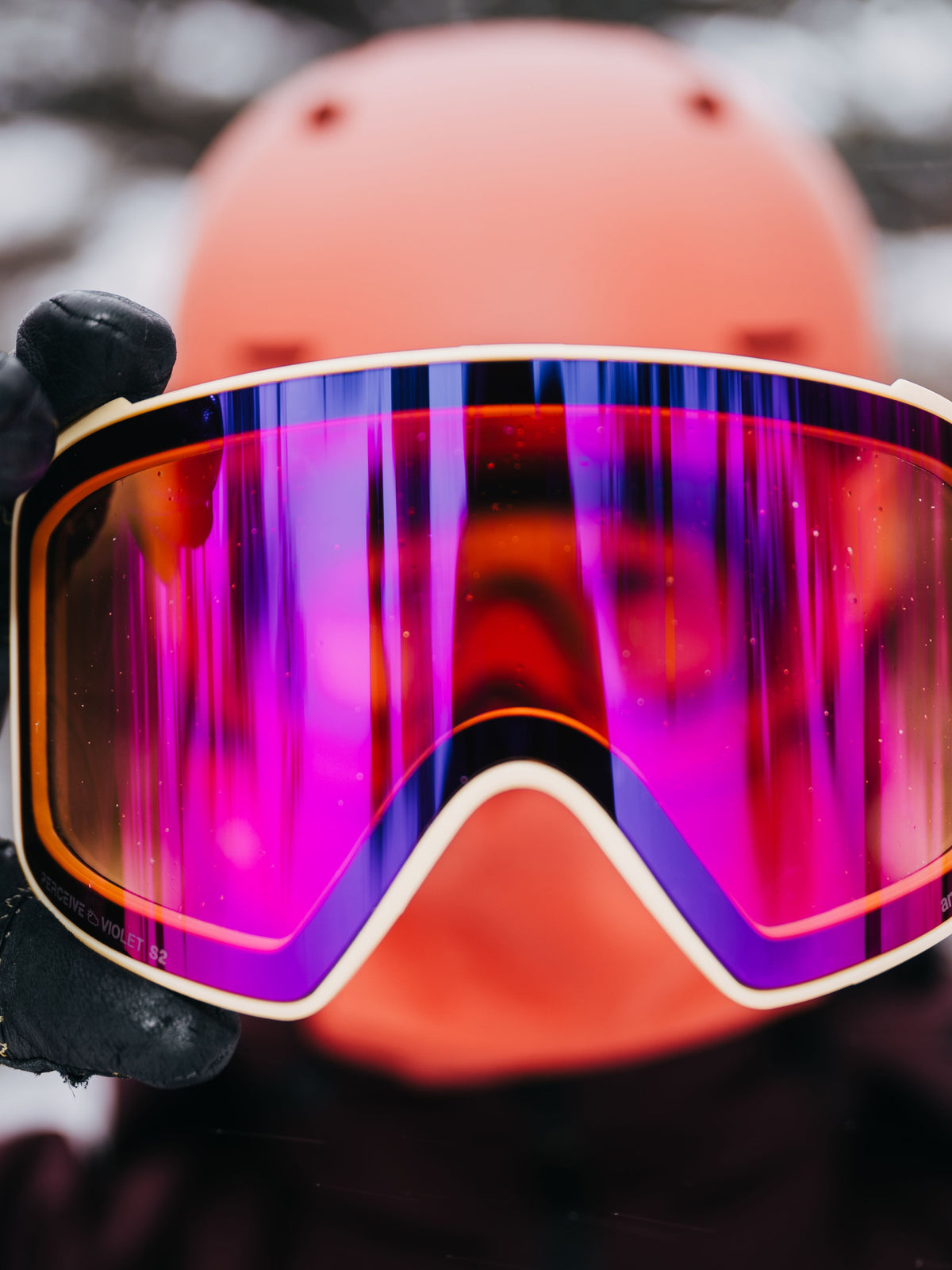 Close-up of a person wearing ski goggles with a large reflective magenta and purple lens, held in front of a blurred orange helmet and snowy outdoor background.