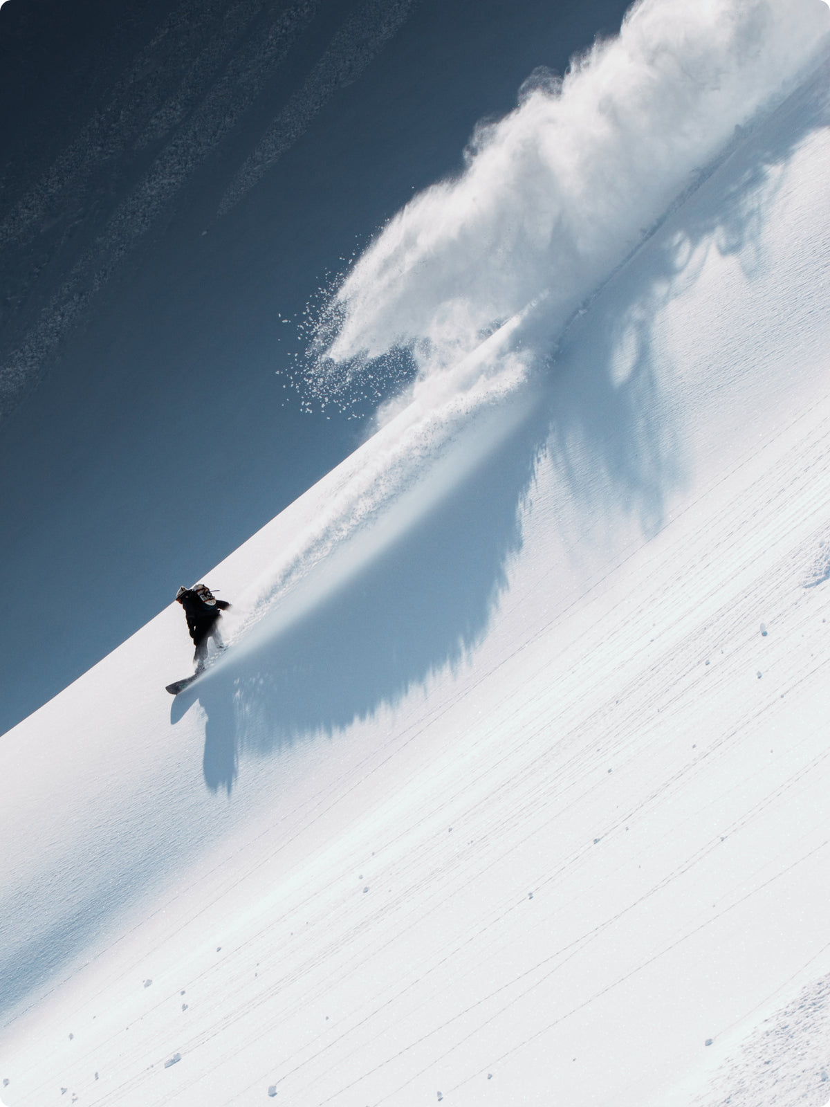 A snowboarder carving through deep powder snow on a steep slope, kicking up a large spray of snow under a deep blue sky.