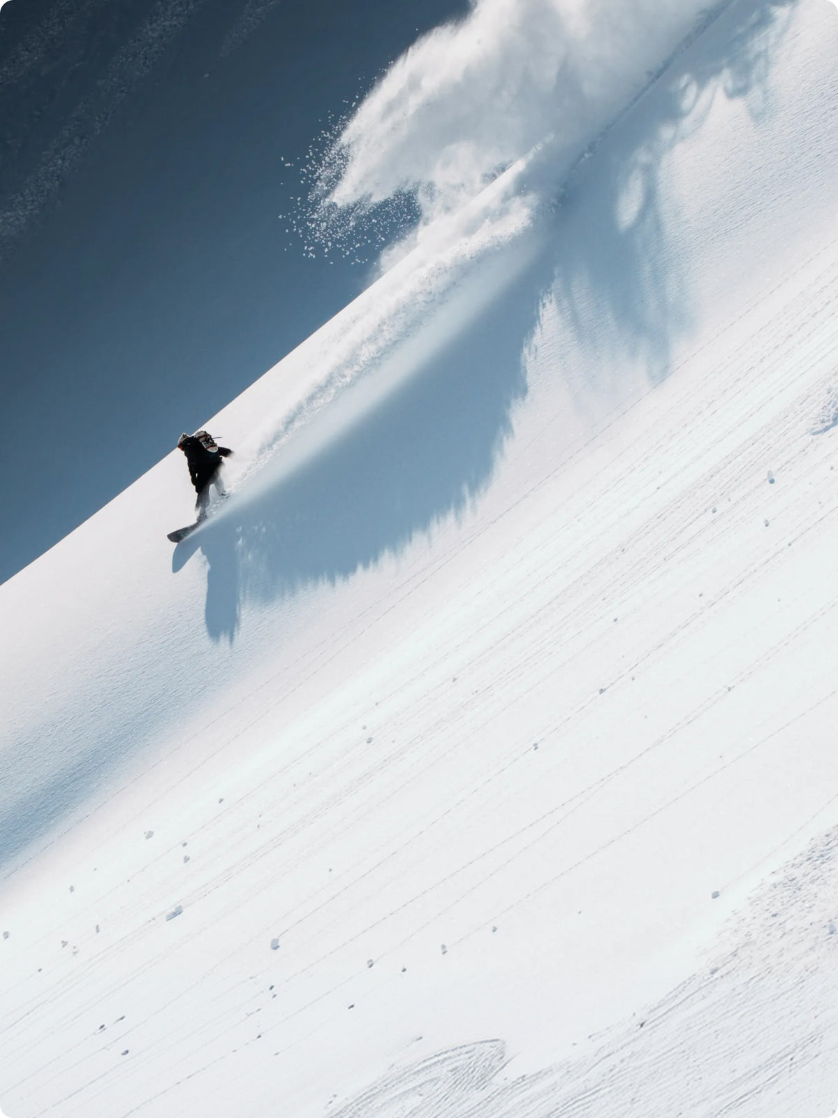 A snowboarder carving through deep powder snow on a steep slope, leaving a long shadow and kicking up a spray of snow under a clear blue sky.