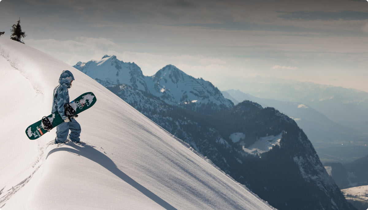 A snowboarder in a light blue cloudy pattern jacket and light blue pants walks down a snowy mountain slope while carrying a green snowboard with white graphics. Snow-covered mountains and a pale cloudy sky are visible in the background.