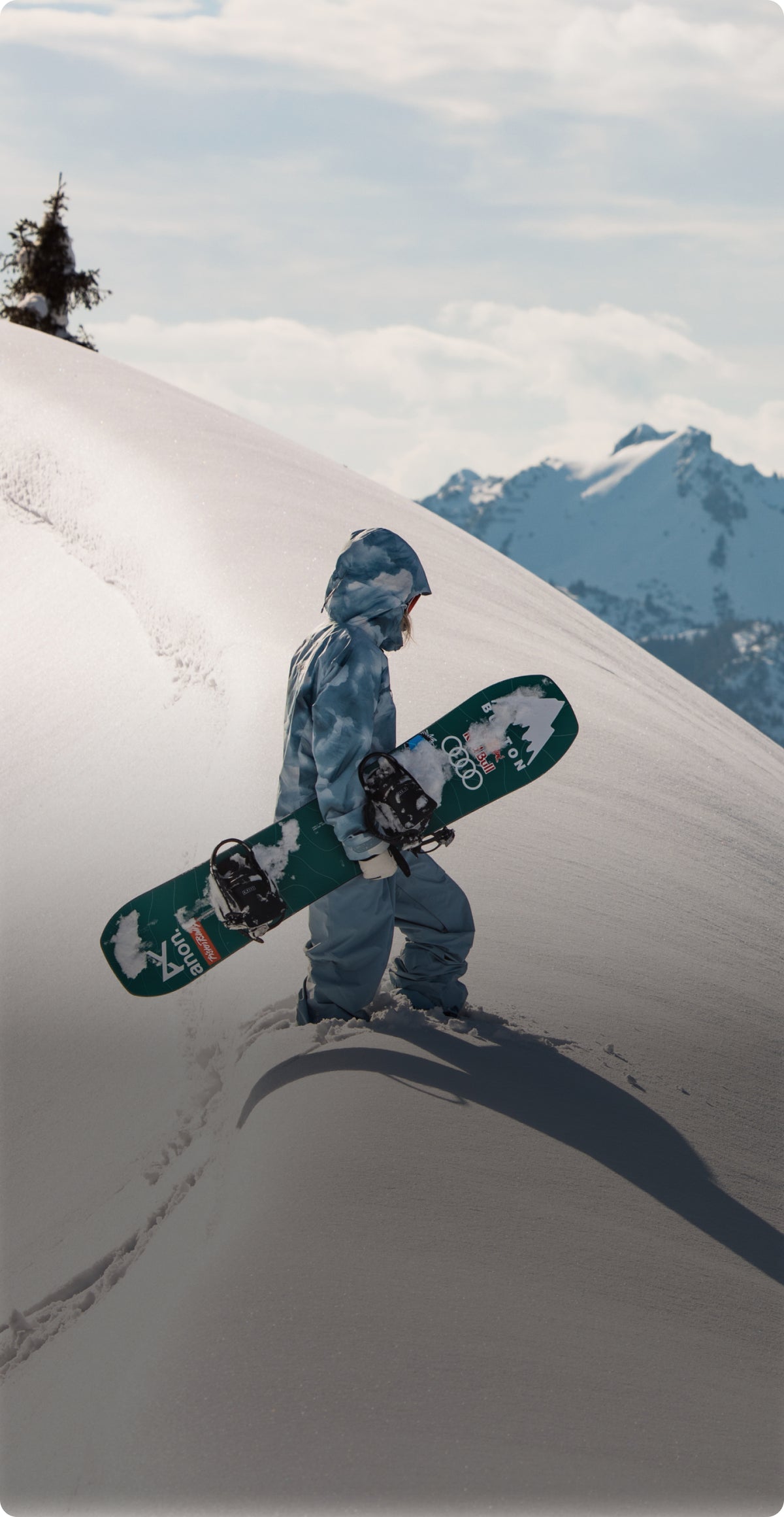 A snowboarder in a light blue cloudy pattern jacket and light blue pants walks down a snowy mountain slope while carrying a green snowboard with white graphics. Snow-covered mountains and a pale cloudy sky are visible in the background.