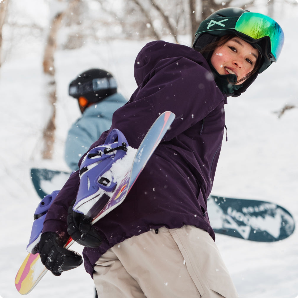 A woman in a purple jacket carrying her snowboard up the hill in the snow.