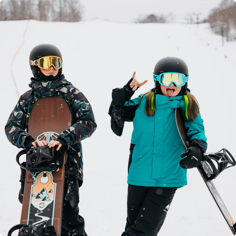 Two snowboarders in winter gear pose in the snow. The person on the left wears a dark camouflage snow jacket, black helmet, gold-tinted goggles, and holds a brown snowboard. The person on the right has a blue jacket and is sticking out their tongue at the camera.