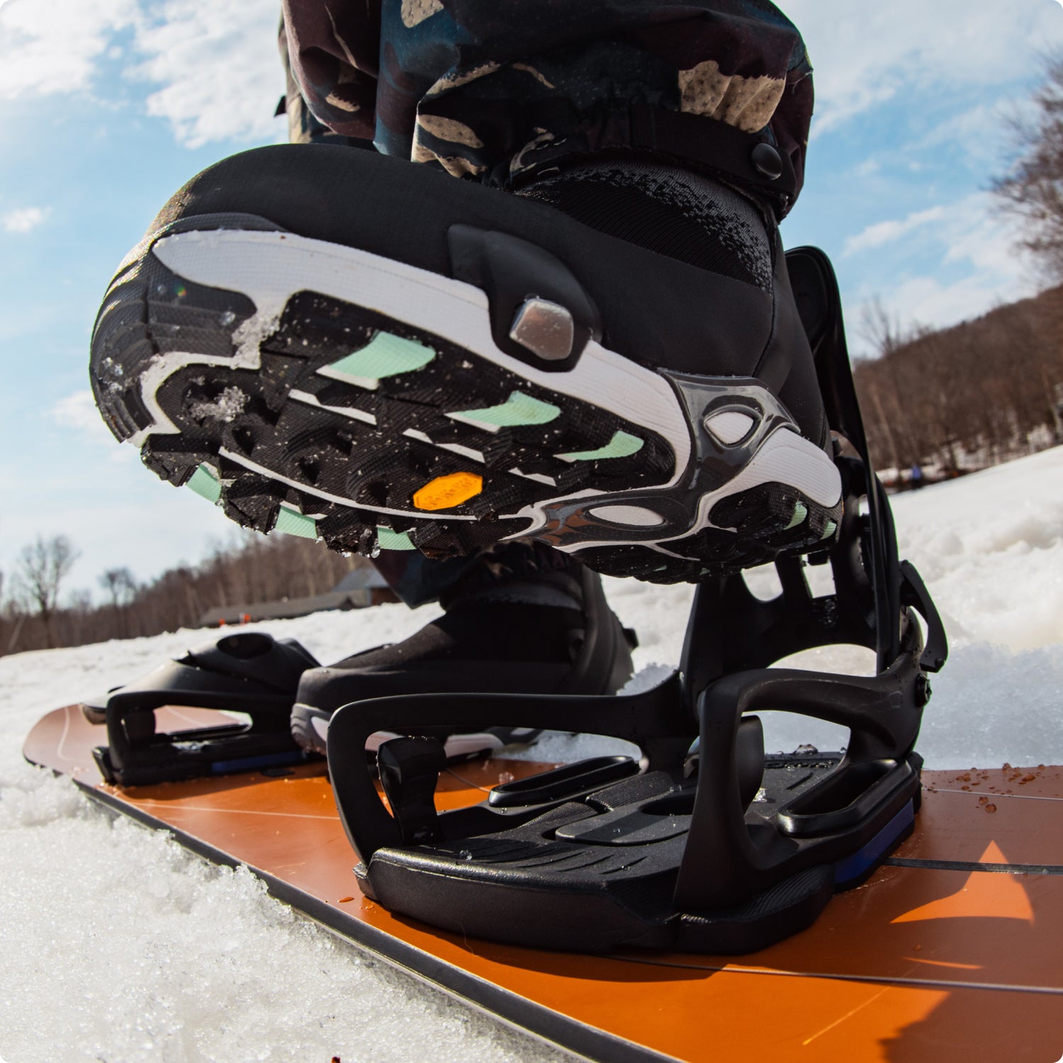 Close-up of a snowboarder's Step On boot and bindings on an orange snowboard in the snow.