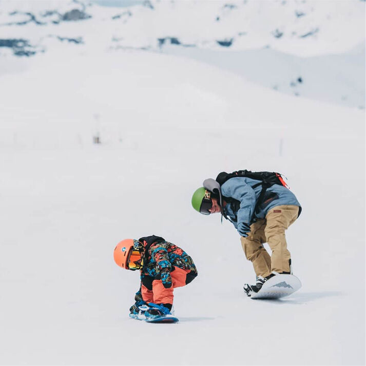 Two snowboarders on a snowy slope with one bending over and the other adjusting their binding.