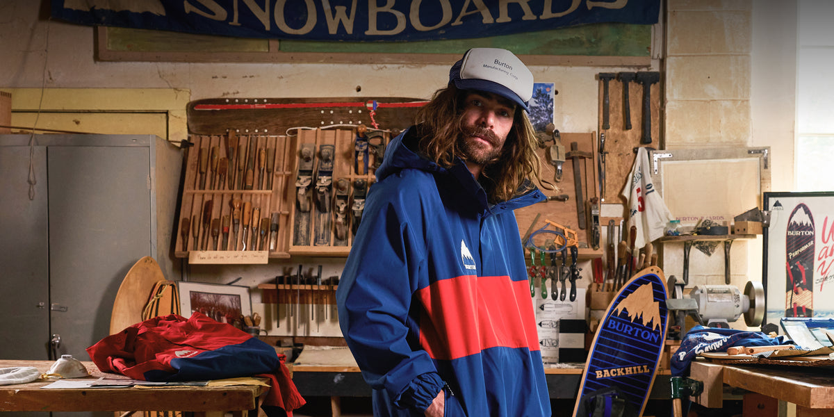 Danny Davis wearing a blue, red, and white color-blocked ski jacket and a white cap, standing in a workshop filled with tools and equipment.