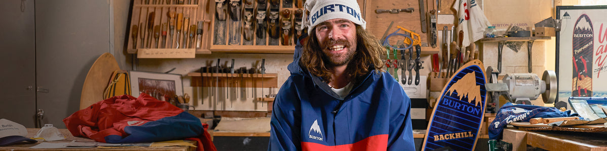 A smiling man wearing a white beanie with 'HISTORIC' printed on it and a blue, red, and white jacket stands in a workshop filled with vintage skis and wooden tools. A blue and yellow 'HISTORIC BACKBIRDS' board is held up on the right.