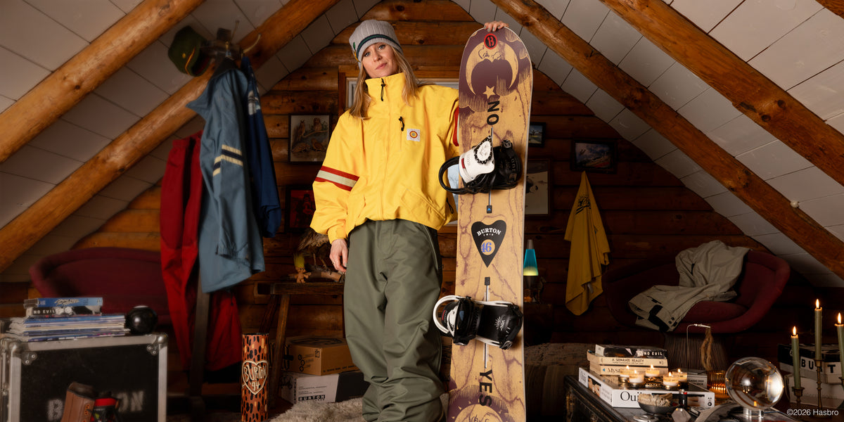 A person stands in a rustic attic room holding a snowboard. The person is wearing a bright yellow jacket, olive green snow pants, and a gray beanie.