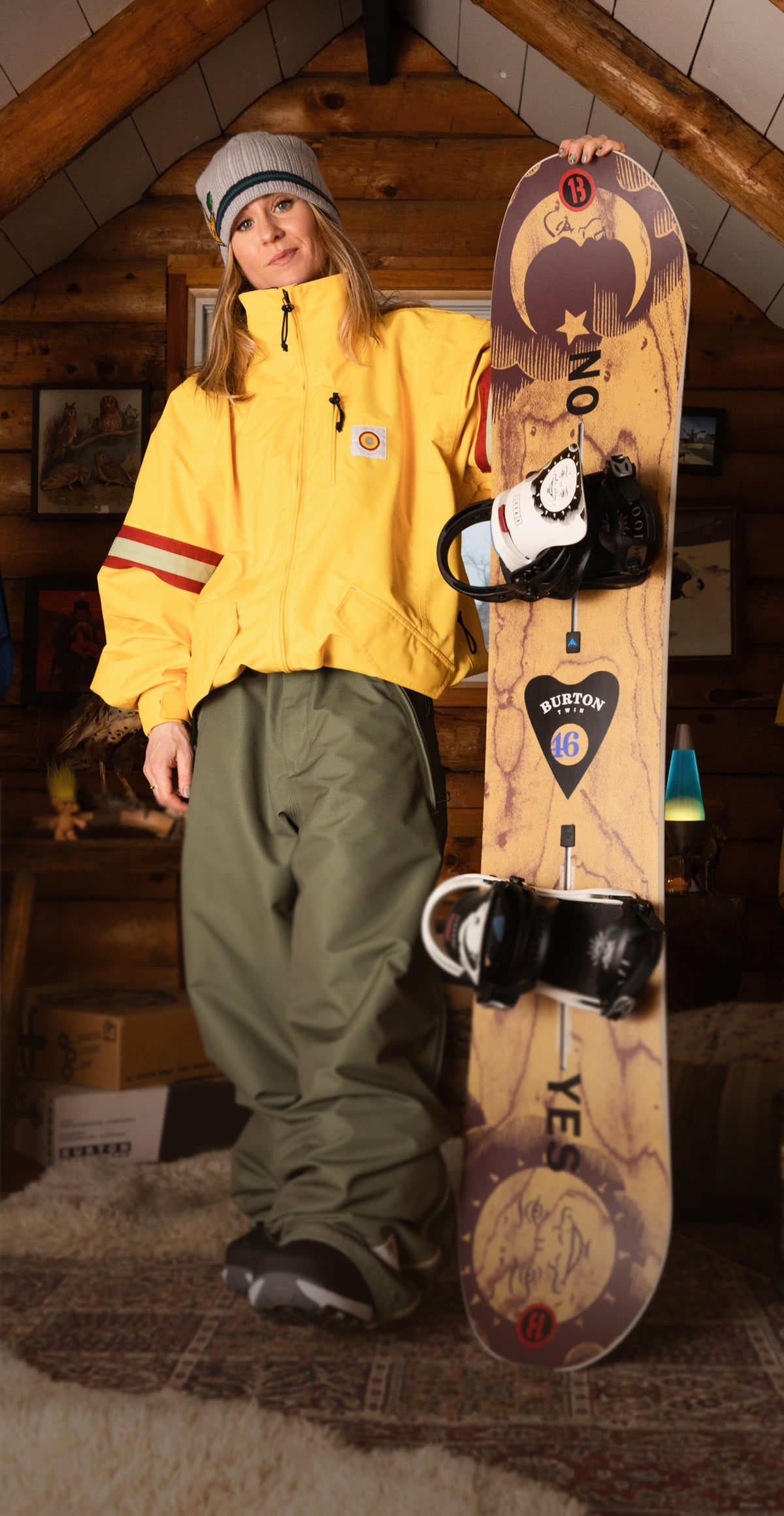 A person stands in a rustic attic room holding a snowboard. The person is wearing a bright yellow jacket, olive green snow pants, and a gray beanie.
