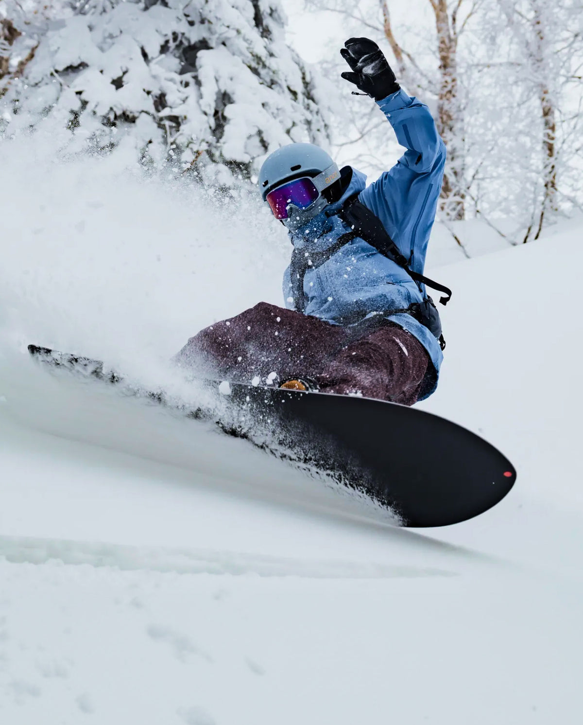 A snowboarder in a blue jacket, light blue helmet, and dark pants carving through deep powder snow, with a black snowboard visible and snowy trees in the background.