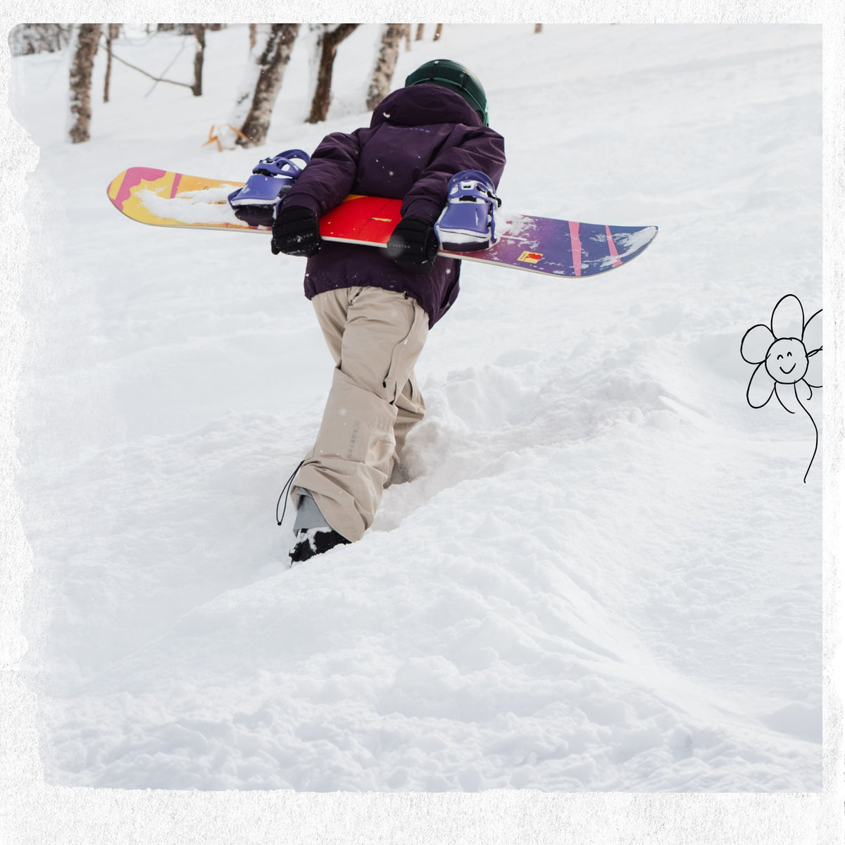 A person in a dark purple winter jacket and beige snow pants carries a colorful snowboard over their shoulder while walking through deep snow outdoors.