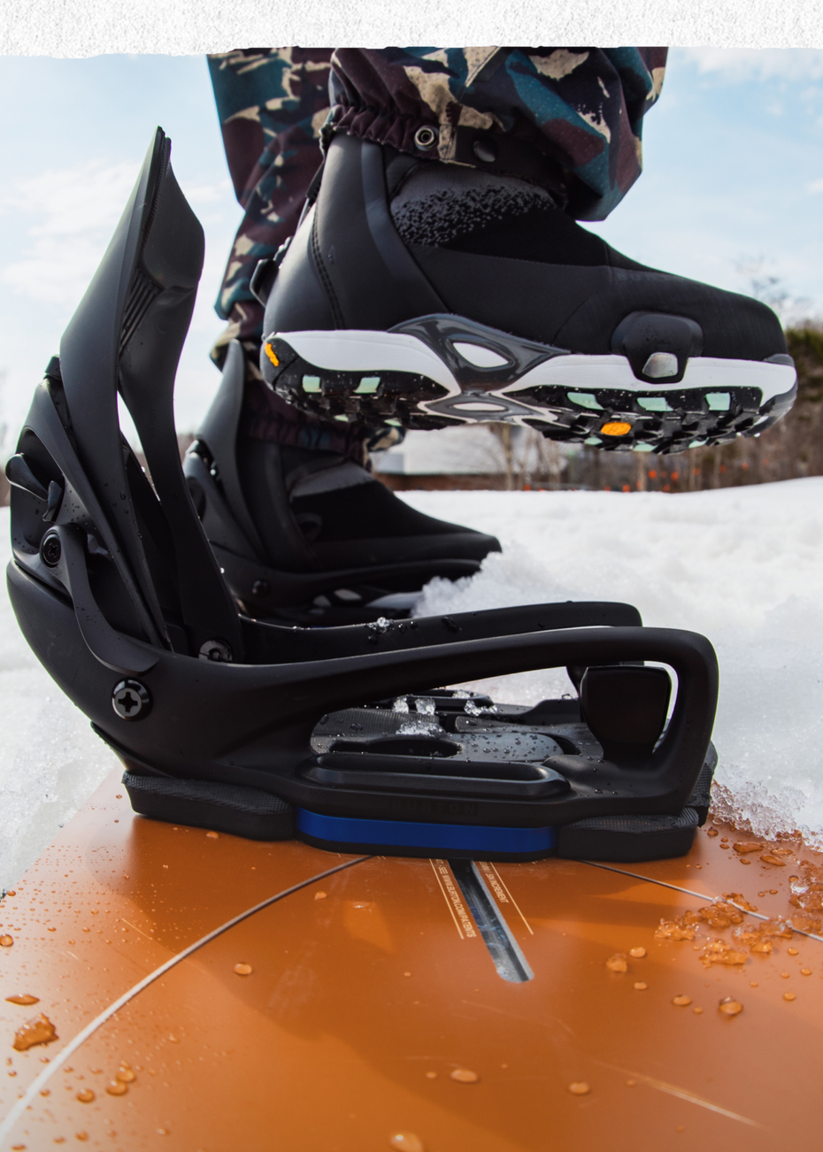 Black snowboard bindings with a blue accent plate, shown on an orange snowboard with snow and a person's boots visible in an outdoor winter setting.