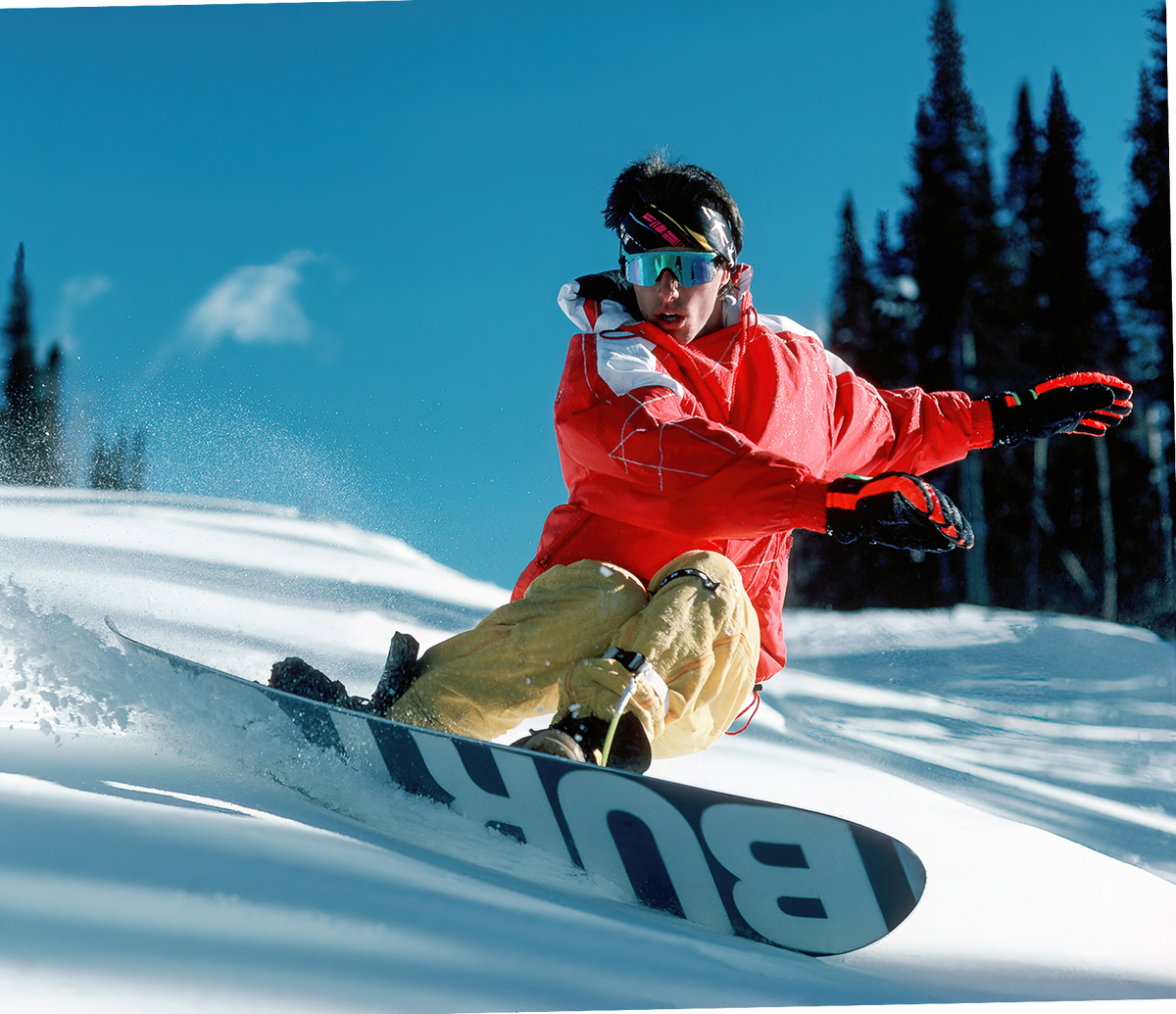 Snowboarder Craig Kelly on snow riding an early Burton Model in a red jacket and yellow pants